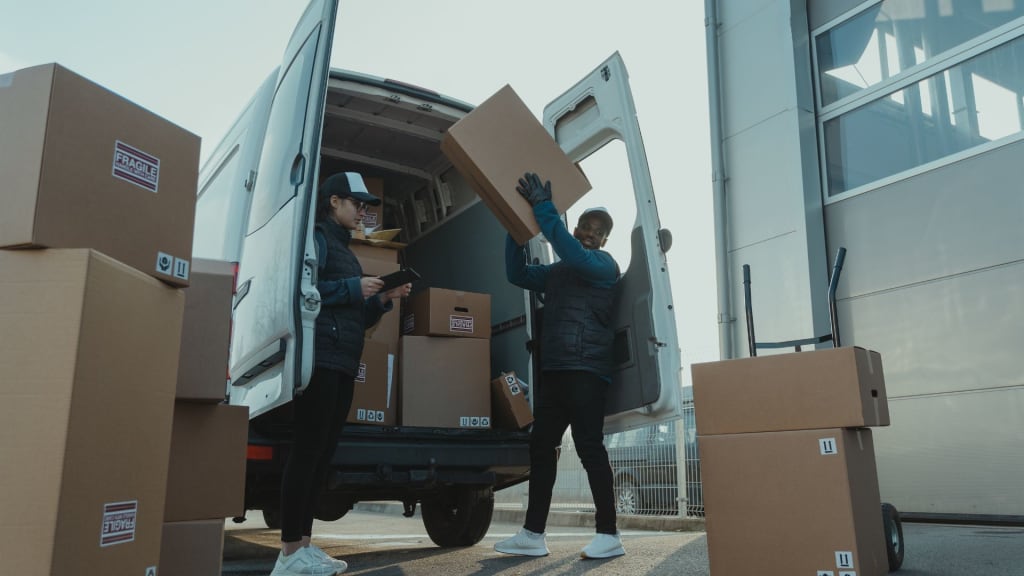 Two workers unload cardboard boxes labeled "fragile" from a white delivery van outside a building, suggesting a wholesale liquor license delivery, with several boxes stacked nearby.
